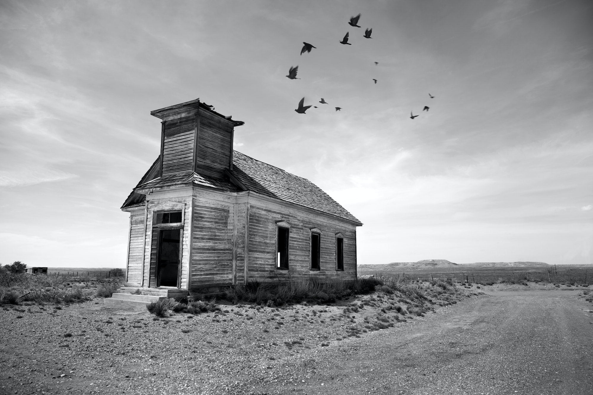 Exterior view of abandoned wooden chapel in remote area, flock of birds in cloudy sky.